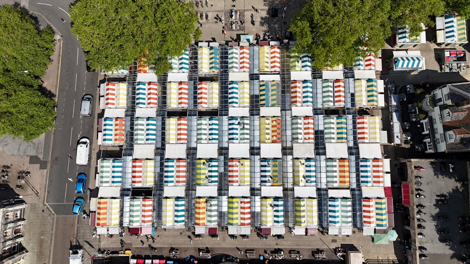 An aerial view captured during the day shows a densely packed outdoor market area with numerous stalls covered by striped awnings in various colors, including blue, red, yellow, and green. The stalls 