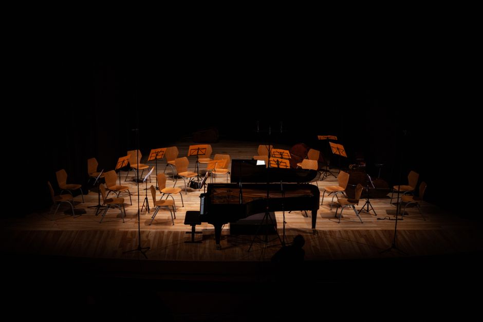 A young woman with long hair, wearing a dark long-sleeved top, is seated at a grand piano in a spacious indoor area with a modern design. The piano, positioned near a wall with a curved white surface,